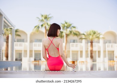 Summer Vacation At Luxury Resort. Back View Of Pretty Young Lady In Elegant Pink Swimwear, Sitting At The Edge Of The Swimming Pool With A Colorful Refreshing Cocktail. Appartments And Palm Tree