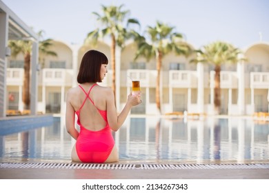 Summer Vacation At Luxury Resort. Back View Of Pretty Young Lady In Elegant Pink Swimwear, Sitting At The Edge Of The Swimming Pool With A Colorful Refreshing Cocktail. Appartments And Palm Tree