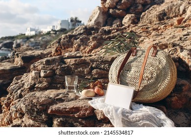 Summer Vacation. Picnic Breakfast On Beach. Straw Basket, Olive Tree Branches. Blank Mockup Card. Glass Of Water, Peach Fruit. Blurred Background With Sea, White Houses. Selective Focus. Mallorca.