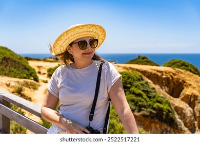 Summer vacation in Portugal. Portrait of woman in beige t-shirt, sunglasses and sun hat standing on wooden pier on cliffs during sightseeing Algarve beaches on beautiful sunny day.
 - Powered by Shutterstock