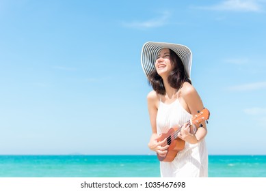 Summer Vacation. Smelling Asian Women Relaxing And Playing An Ukulele On The Beach, So Happy And Luxury In Holiday Summer, Outdoors Blue Sky Background. Travel And Lifestyle Concept.