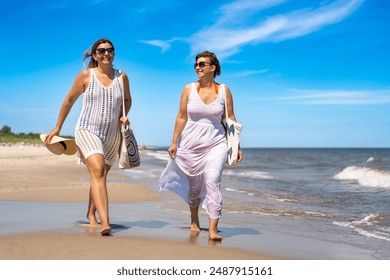 Summer vacation. Two mid-adult smiling women wearing swimsuits and white summer dresses with beach bags walking to beach by sea while talking. Walk on beach on the Baltic Sea. Front view - Powered by Shutterstock