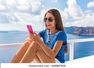 Summer Vacation Woman Using Cellphone Texting Sms Message Outside On Balcony Terrace Of Luxury Hotel With Mediterranean Sea In Oia, Santorini. Europe Happy Young Girl Relaxing In Sun On Mobile Phone.