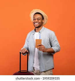 Summer vacation. Young black man in casual clothes with suitcase, passport and tickets over orange studio background. African American guy traveling on weekend, ready for plane flight