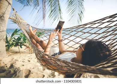 Summer vacations concept, Happy woman with white bikini, hat and sunglass using mobile phone in hammock on tropical beach at sunset, Koh mak, Thailand