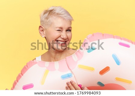 Summer, vacations, seasons and lifestyle. Isolated image of happy funny middle aged woman with short blonde hair fooling around on beach by sea, posing against yellow background with swimming circle