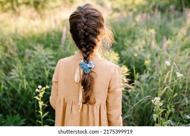 Summer vibes. The feeling of summer. Little girl having french plait hairstyle wearing linen mustard dress walking on the green fields. People from behind