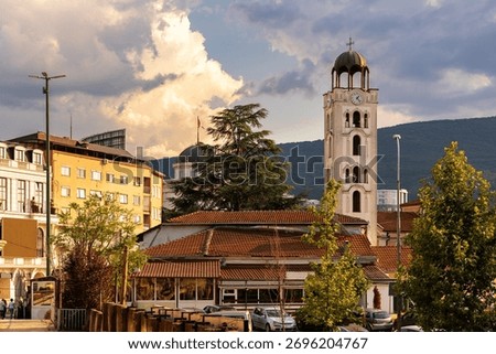 Summer view of Old Bazaar district in Skopje, North Macedonia, with bell tower of St. Clement of Ohrid Cathedral rising above red rooftops of buildings, and green slopes of Mount Vodno in background