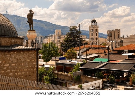 Summer view of Old Bazaar district in Skopje, North Macedonia, with bell tower of St. Clement of Ohrid Cathedral rising above red rooftops of buildings, and green slopes of Mount Vodno in background
