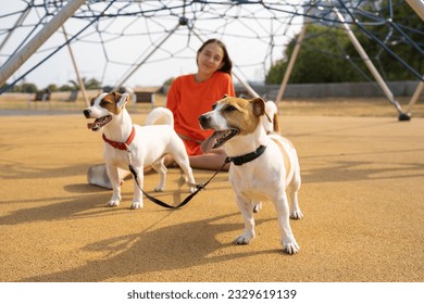 Summer walk with two dog. Teen girl in orange clothes sitting playground with her small cute white Jack Russell terriers pets with double leash. Summer in big cityю girl in the background out of focus - Powered by Shutterstock