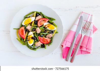 Summer Warm Salad With Cooked Green Beans, Tuna, Tomatoes, Boiled Eggs And Sauce Balsamico Glassa In White Plate With Knife And Fork On Wooden Background. Healthy Eating Concept. Top View.