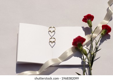 Summer wedding mockup. Blank wedding book flowers on a white table. Wedding rings are placed on the book and the sunlight creates a heart-shaped shadow. Carnations and ribbon.