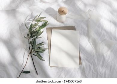 Summer wedding stationery, birthday mock-up scene. Blank greeting card with envelope, silk ribbon and olive tree branches. White linen textile background in sunlight, long shadows. Flat lay, top view. - Powered by Shutterstock