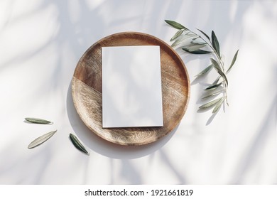 Summer wedding stationery mock-up scene. Blank greeting card, wooden plate, olive tree leaves and branches in sunlight. White table  background with palm shadows. Feminine flat lay, top view. - Shutterstock ID 1921661819
