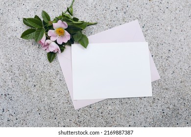 Summer Wedding Stationery Mock-up Scene. Horizontal Blank Greeting Card, Invitation, Pink Envelope And Blooming Dog Rose Flowers On Textured Terrazzo Background. Flat Lay, Top View. No People.