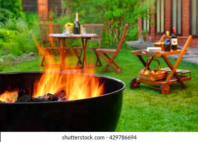 Summer Weekend BBQ Scene On The Backyard. Flaming Charcoal Grill Close Up. Outdoor Wooden Furniture  On The Blurred Background.