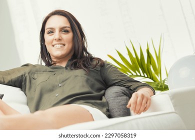 In summer, a woman in white shorts and a military green shirt is barefoot, sitting and lying down on a couch in her living room. The colors are bright, and she's happy, smiling, relaxed, and self-conf
