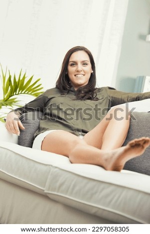 In summer, a woman in white shorts and a military green shirt is barefoot, sitting and lying down on a couch in her living room. The colors are bright, and she's happy, smiling, relaxed, and self-conf