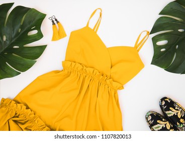 Summer women's fashion flat lay clothing and accessories: yellow dress, yellow earrings with a brush, colorful mules and palm leaves on a white background. Summer fashion shopping concept. 