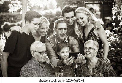 Summertime, A Family With Three Generations Having Fun Around A Table In The Garden Sharing A Meal. The Whole Guest Having Fun Watching A Video On A Phone. Black And White