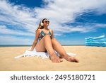 Summertime with music. Beautiful young woman in colorful swimsuit and sunglasses listening to music in headphones while sitting on towel on white sandy beach.