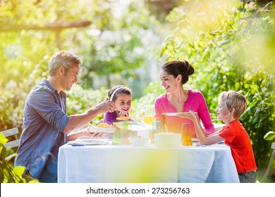 Summertime, Nice Family, Daddy, Mom And Their Two Kids Sitting At A Table, Eating Lunch In The Garden Enjoying A Sunny Day