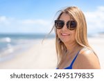 Summertime on beach. Portrait of middle-aged beautiful smiling woman with long blonde hair in blue bikini and sunglasses against background of sandy beach and sea on sunny day. 