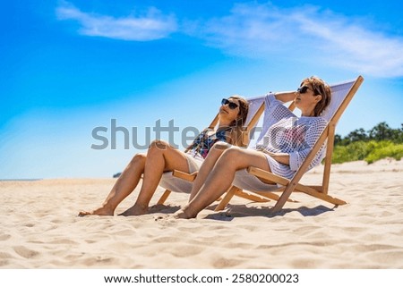 Summertime on beach. Two beautiful middle aged women sitting on deckchairs on white sandy beach