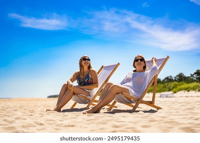 Summertime on beach. Two young women of European beauty dressed in beach wear looking at sky while sitting on deckchairs on beach with white sand on beautiful sunny day. Relaxing on beach. - Powered by Shutterstock