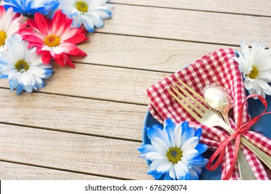 Summertime Picnic Table Place Setting With Red White And Blue Flowers, Plate, Napkin, Fork And Spoon On Rustic Wood Board Background With Blank Room Or Space In Center For Copy, Text, Or Your Words.