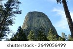 The summit of Devils Tower rises above surrounding pine trees under a partly cloudy sky in Wyoming