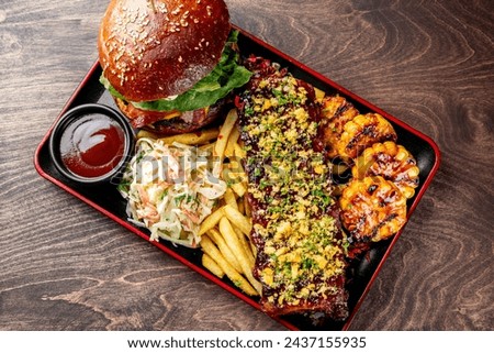 A sumptuous meal awaits: a gourmet burger, ribs, fries, coleslaw, and grilled corn on a dark wooden table.