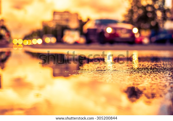 Sun after the rain in the city, view of the\
approaching car with a level of puddles on the pavement. Image in\
the yellow-purple toning