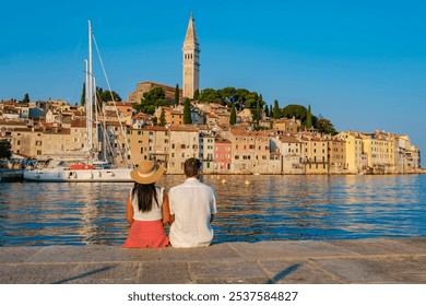 As the sun begins to set, a couple sits by the calm waters in Rovinj, Croatia, admiring the vibrant buildings and reflecting on the day's adventures. A sailboat adds to the picturesque backdrop. - Powered by Shutterstock