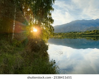 The sun behind the trees, shining through the leaves, illuminates the lake's shore in the glow of sunset. The water reflects the bright light - Powered by Shutterstock