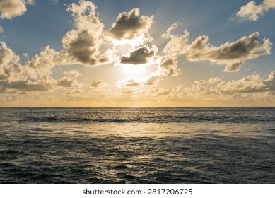 Sun breaks through dramatic clouds above calm sea, casting shimmering reflections on the water at dusk in Seychelles.