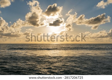 Sun breaks through dramatic clouds above calm sea, casting shimmering reflections on the water at dusk in Seychelles.