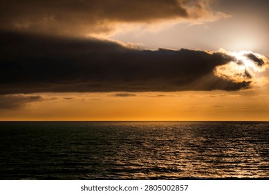 The sun breaks through heavy clouds at sunset, casting a warm golden glow over the dark, rippling ocean surface