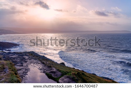 The sun breaks through storm clouds and reflects off waves breaking on the shore, the seaside village of Woolacombe just visible in the distance