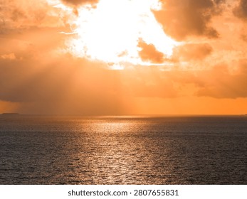 Sun breaks through thick clouds over the Indian Ocean near Dhigurah Island, casting a molten orange glow on calm rippling water, with low land faint on the horizon.