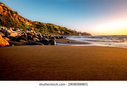 The sun casts a warm glow on the sandy beach and rugged cliffs of Palos Verdes Estates, with the Pacific Ocean gently lapping the shore