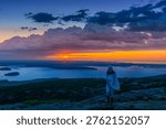Sun dips behind distant waters, painting sky in fiery orange. Lone figure stands on rocky ridge, wrapped in soft, flowing blanket. Dramatic clouds swirl above, casting deep blue shadows