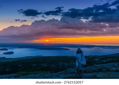 Sun dips behind distant waters, painting sky in fiery orange. Lone figure stands on rocky ridge, wrapped in soft, flowing blanket. Dramatic clouds swirl above, casting deep blue shadows