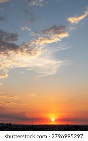 Sun dips below distant skyline, painting sky in gold and orange. Clouds streak across blue, softly illuminated by fading light. Cityscape lies silhouetted, quiet under the setting sun's glow