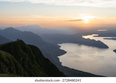 The sun dips below the horizon, casting a warm golden glow over the iconic Lake Lucerne and the surrounding mountains. Silhouetted figures stand atop Niederbauen Kulm, taking in the breathtaking panoramic view of the lake and the Swiss Alps. The scene evokes a sense of awe and tranquility, capturing a perfect moment of natural beauty.