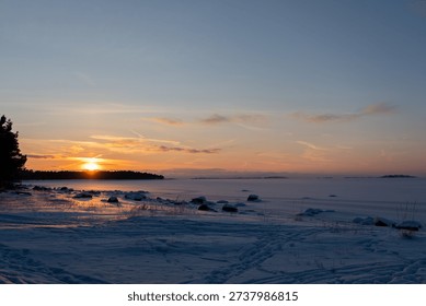 The sun dips below the horizon casting warm hues across the sky over a tranquil frozen seascape with rocky, snow-laden coastline