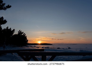 The sun dips below the horizon casting warm hues across the sky over a snow-covered landscape and icy water