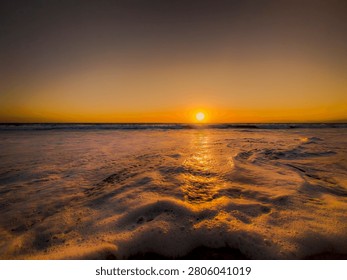The sun dips below the horizon, casting a warm, golden glow across the sea and illuminating the foamy waves