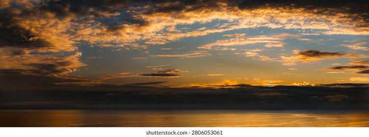 The sun dips below the horizon, casting a warm golden glow on the vast ocean and illuminating the dramatic clouds above