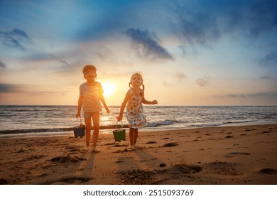 As the sun dips below the horizon, two kids walk on the beach, each holding a bucket, with calm waves in the background